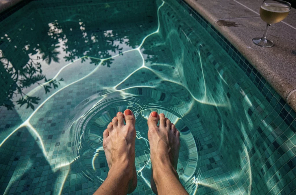Gros plan de pieds dans l'eau d'une piscine éclairée la nuit avec des reflets turquoise et un motif en mosaïque visible
